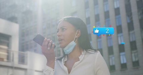 Young professional speaking into smartphone with lowered mask in sunlit downtown