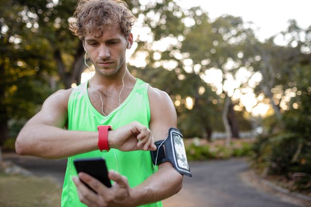 Jogger in green tank top checking his smartwatch while listening to music on his mobile phone in a park. Ideal for use in fitness, technology, and healthy lifestyle promotions. Suitable for illustrating outdoor exercise routines, wearable technology, and active living.