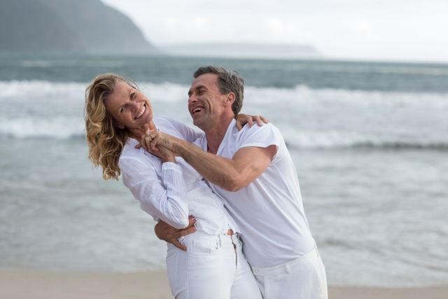 Mature couple laughing and enjoying time together at the beach. Perfect for use in advertisements for travel agencies, vacation packages, retirement planning, and lifestyle blogs focusing on relationships and outdoor activities.