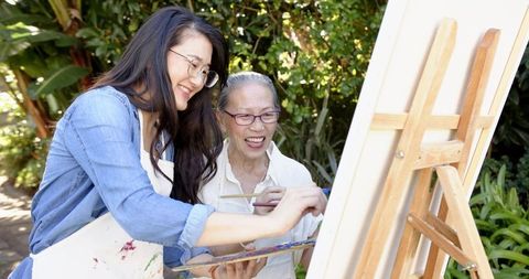 Asian senior and younger woman painting together outdoors teaching and enjoying art