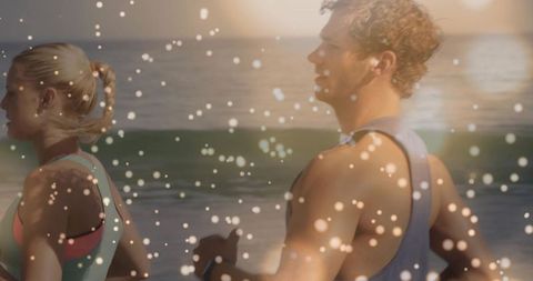 Fit couple jogging along ocean shore at golden hour with bokeh light overlay