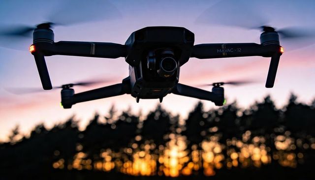 Hovering Black Quadcopter Drone with Camera Gimbal Over Forest Silhouette at Sunset