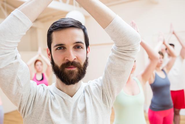Man practicing tree pose in a fitness studio with a group of people in the background. Ideal for use in content related to yoga, fitness, wellness, group exercise classes, and healthy living.