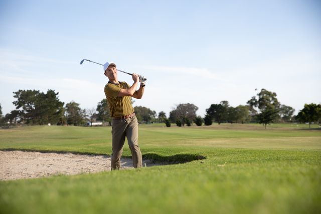 Caucasian male golfer practicing his swing on a sunny day at a golf course. He is wearing a cap and golf clothes, focusing on hitting the golf ball. Ideal for use in content related to sports, outdoor activities, healthy lifestyles, hobbies, and leisure.