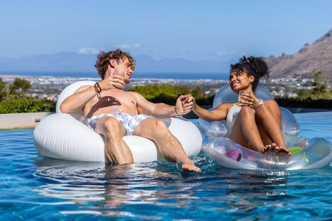 Young couple floating and holding hands in infinity pool with coastal view and drinks
