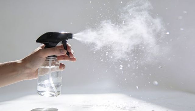 Closeup left hand spraying fine mist from clear trigger bottle with droplets and nozzle