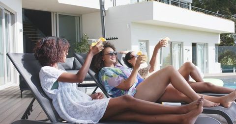 African american friends relaxing poolside at modern villa toasting with citrus cocktails