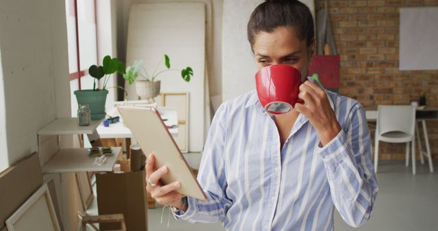 This image depicts a woman drinking coffee while working with a clipboard in an art studio. She appears focused and immersed in her work. Ideal for illustrating concepts of creativity, concentration, and casual work environments. Suitable for use in articles and advertisements related to art, productivity, and working from home.