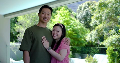 Asian couple embracing on modern balcony smiling, showing smartwatch and pink headband