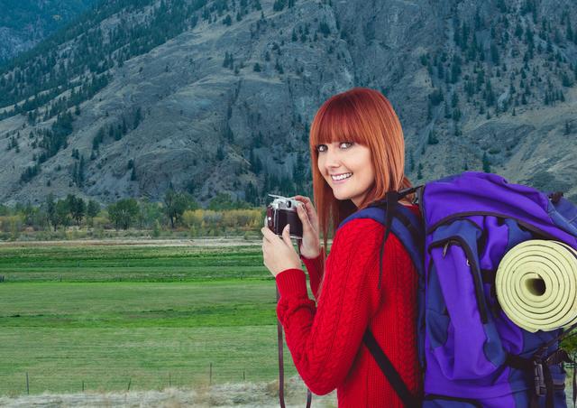 This image shows a redheaded woman wearing a red sweater and carrying a large purple backpack in a mountainous landscape. She is smiling and holding a camera, capturing the scenic environment around her. The green valley and towering mountains suggest a serene and adventurous travel experience. This image can be used for promoting outdoor activities, travel blogs, adventure gear, tourism websites, and photography workshops.
