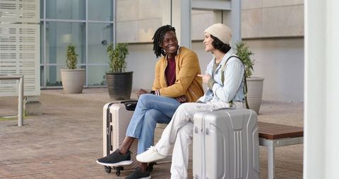 Smiling diverse travelers sitting with luggage on urban bench, talking and planning