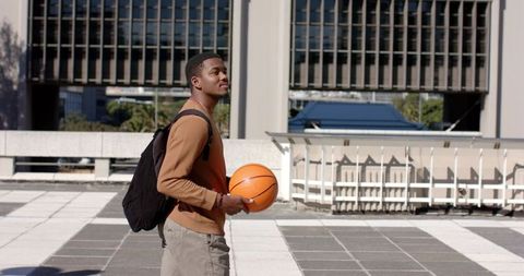 African american student walking across campus plaza holding basketball and backpack