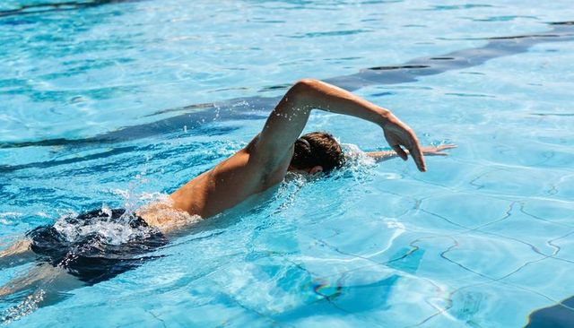 Male swimmer performing freestyle stroke in sunlit outdoor pool, water splash motion