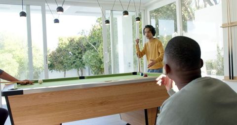 Multicultural friends playing pool in sunlit modern game room with glass doors
