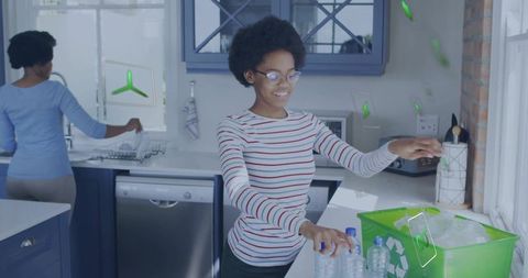 Teen placing plastic bottles into green recycling bin while family member washes dishes