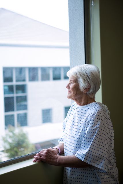 Senior patient standing by hospital window, looking outside with sense of contemplation. Ideal for use in health care promotions, senior care marketing, medical brochures, and articles on aging or hospital environments.