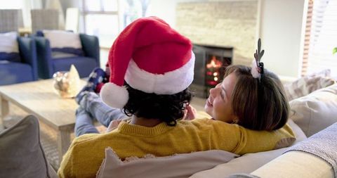 Couple cuddling on sofa wearing Santa hat and reindeer antlers by cozy glowing fireplace