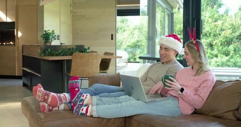 Couple relaxing on leather sectional wearing santa hat and reindeer antlers using laptop