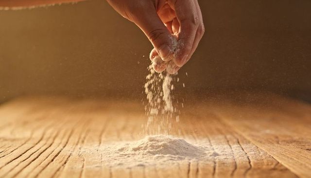 Hand sprinkling flour over rustic wooden counter dusting warm light baking moment