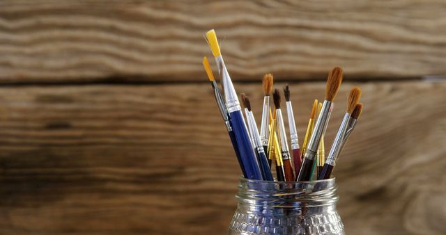 An assortment of paintbrushes is placed in a glass jar against a rustic wooden background. The brushes have varied shapes, sizes, and colors, highlighting a creative and artistic vibe. This image can be used for art supply advertisements, creative workshops promotions, art studio decor, and blogs about painting and crafts.