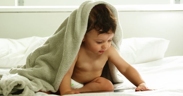 Adorable baby sitting on bed wrapped in a blanket, attentively engaged with an object. Scene captures innocence and curiosity. Suitable for family, parenting, early childhood education, home comfort, or cozy home-themed content.