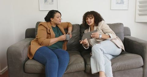African American Women Sharing Tablet and Coffee Discussing Ideas on Cozy Sofa