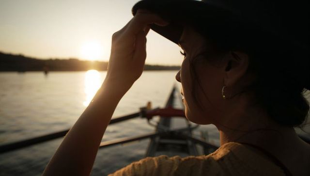 Asian rower holding hat brim while gazing at sunset over lake in rowing shell