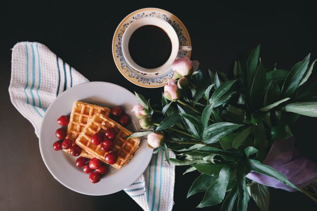 Perfect for food blogs and websites, this image captures a tasteful breakfast setup featuring golden waffles with fresh cherries beside a cup of coffee and a bouquet of flowers. Ideal for promoting breakfast recipes, café menus, or lifestyle content. The dark background highlights the vivid colors of the breakfast items, making it visually appealing.