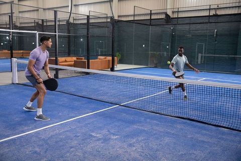 Two diverse men playing padel on indoor blue court at net, mid-swing competitive match