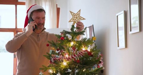 Man in Santa Hat Decorations While Talking on Phone by Christmas Tree