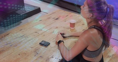 Female athlete sitting on wooden box in gym, coffee cup and phone nearby, colorful lights