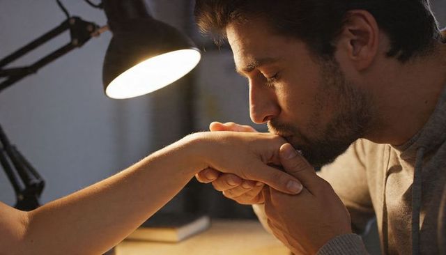 Bearded man kissing partner hand under warm lamp glow, intimate tender moment
