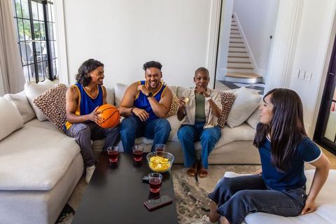 Cheering friends watching basketball at home sitting on couch holding basketball and snacks