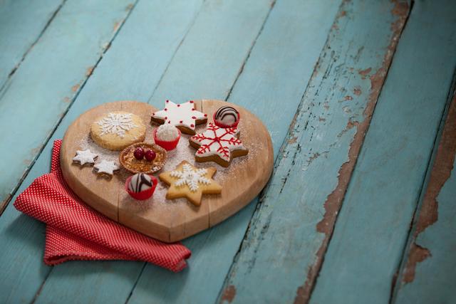 Beautifully decorated Christmas gingerbread cookies with intricate icing on a heart-shaped wooden board, set against a rustic blue wooden table. Perfect for holiday greeting cards, festive recipes, Christmas blog content, and food and lifestyle magazines.