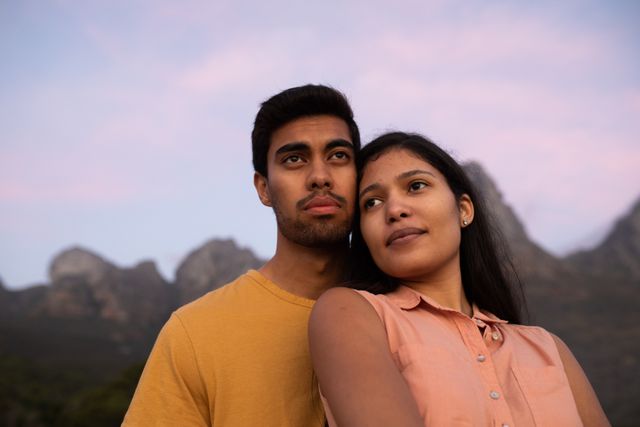 Biracial couple standing together in nature, embracing and admiring the scenic mountain view at sunset. Ideal for use in campaigns promoting love, diversity, relationships, outdoor activities, and travel. Perfect for illustrating themes of togetherness, relaxation, and natural beauty.