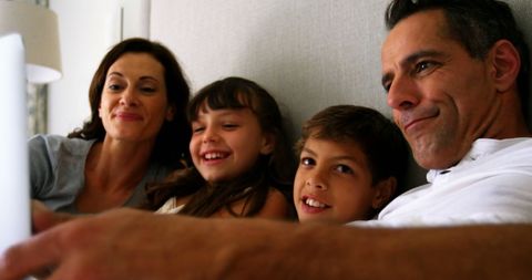 family of four gathers on sofa, engaging with digital device, representing modern family bonding. suitable for concepts related to family joy, technology, home life, happiness, digital entertainment, and intergenerational communication. use for parenting blogs, tech advertisements, or family-oriented content.