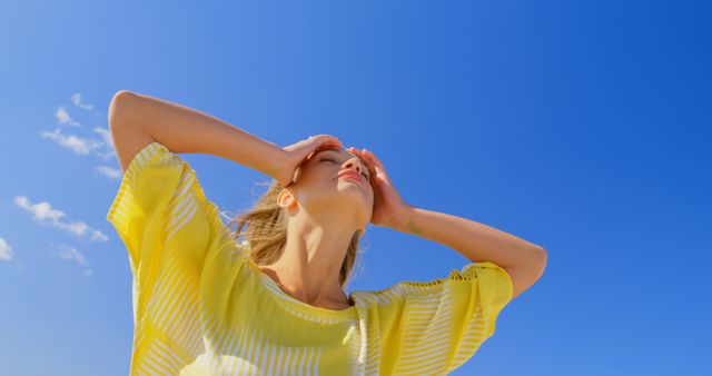Woman with hands on head, standing under bright blue sky, enjoying warm sunny day. Ideal for concepts of summer relaxation, outdoor lifestyle, and carefree nature moments. Perfect for advertisements, travel promotions, and lifestyle blogs.