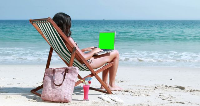 A young woman sits on a beach chair working on a laptop with a green screen, with copy space. Her relaxed posture contrasts with the productivity implied by her remote work setup against the serene beach backdrop.