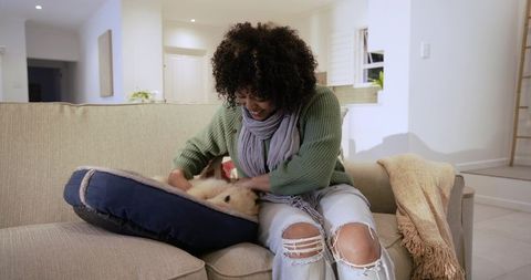 Smiling woman petting puppy on navy dog bed while relaxing on beige sofa