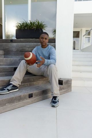 Teenage african american male sitting on modern patio steps holding football