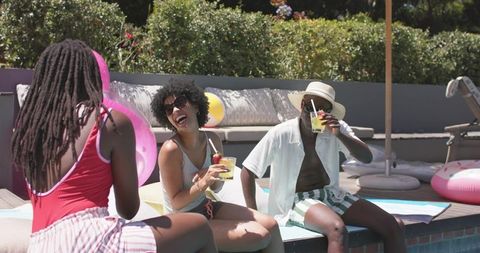African american friends laughing and sipping cocktails by backyard pool on warm sunny day