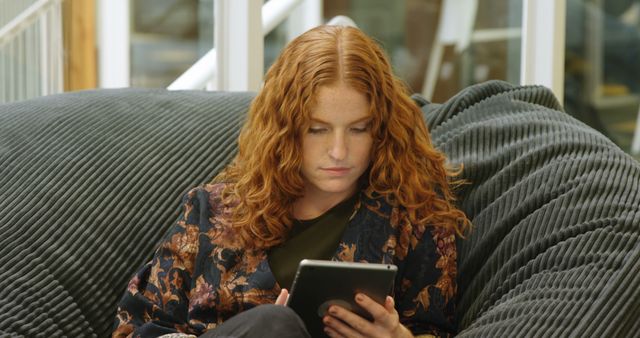 Young woman with red hair is relaxing on a comfortable couch while using a tablet. Perfect for lifestyle or technology themes, home leisure concept, or articles about young adults. Can be used in content related to relaxation, digital entertainment, or modern home living.