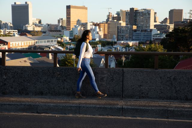 Biracial woman walking along an elevated road with a cityscape in the background. She is wearing a backpack and casual outfit, looking at the city. Ideal for use in urban lifestyle, travel, and city exploration themes.