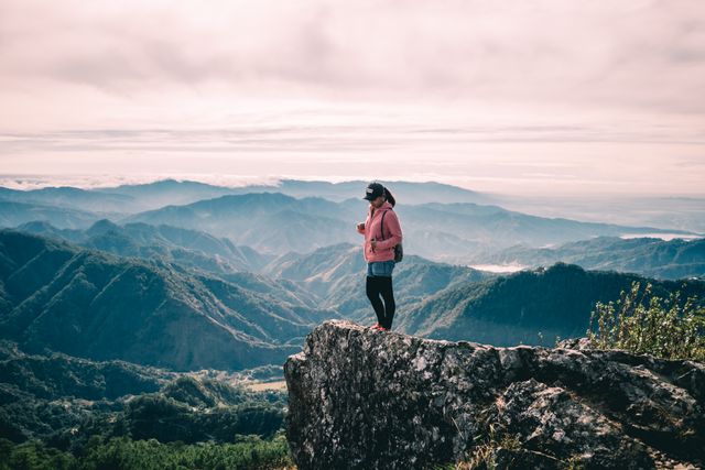 Female hiker standing on edge of mountain cliff, admiring scenic valley below during foggy day. Scene perfect for portraying themes of adventure, exploration, travel, solitude, and the beauty of nature. Ideal for use in travel and adventure blogs, outdoor activity advertisements, inspirational posters, and nature photography features.