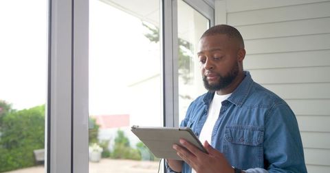 African american man standing in sunroom using tablet with folio case, focused and calm