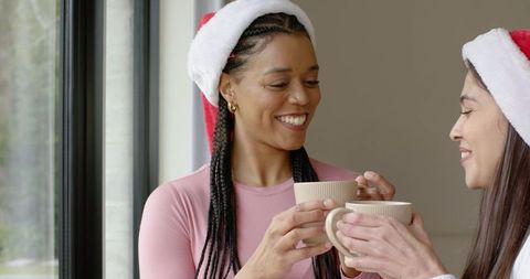 Multicultural women clinking ribbed mugs while wearing santa hats, sharing warm holiday toast