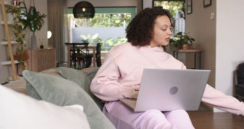 African american woman working on laptop on sofa in cozy sunlit living room with plants