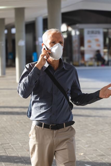 Caucasian man wearing face mask talking on smartphone while walking in city streets during daytime. Useful for topics related to urban life, pandemic safety measures, communication, and public health awareness.