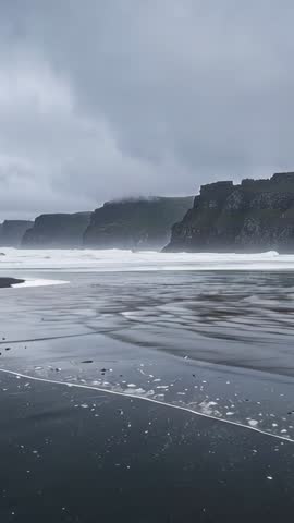 Vertical video: rolling waves on black sand beach reflecting volcanic cliffs