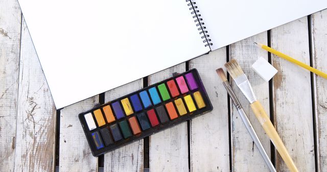 A set of watercolor paints and brushes lie next to a blank sketchbook on a wooden surface, with copy space. The arrangement invites creativity and suggests an artistic project or hobby about to begin.